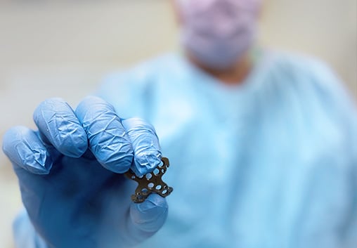 Surgeon in a mask and gown holding up a Summit™ Patella Plate between gloved fingers